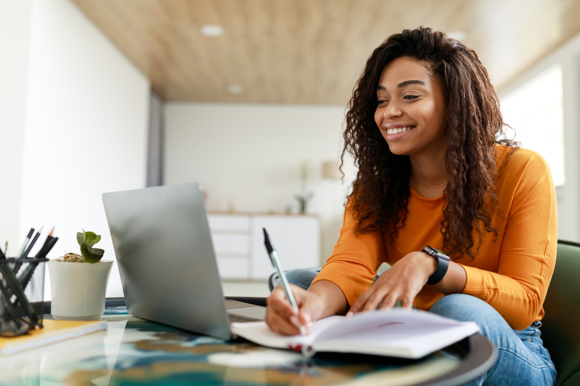 Black woman sitting at desk, using computer writing in notebook